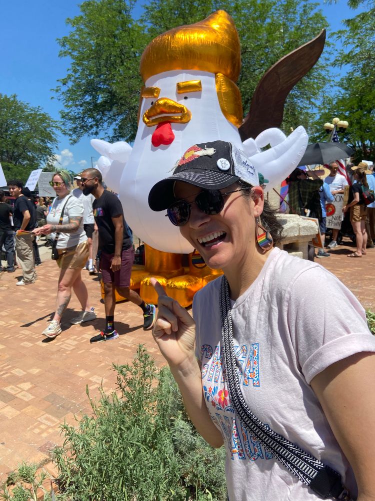 No Kings protest in Pueblo, CO. Sabrina standing in front of, and pointing to, a large inflatable white chicken that comically looks like Trump. 