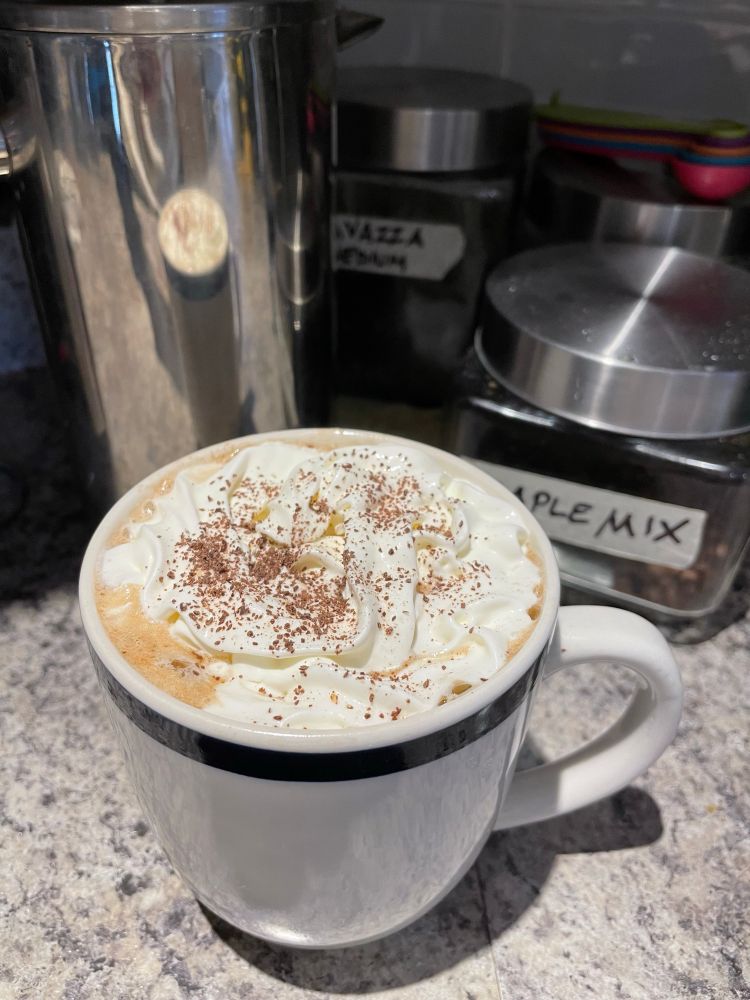 A large white mug with a blue rim sits on a speckled grey stone-look kitchen counter. Behind it are two glass containers with coffee beans and masking tape labels that read Maple mix and Lavazza. Yum!  A stainless steel french press coffee maker shines beside the mug. The mug is filled with coffee, topped with a thick, fluffy cloud of whipped cream and chocolate shavings. This is a coffee lover’s kitchen!