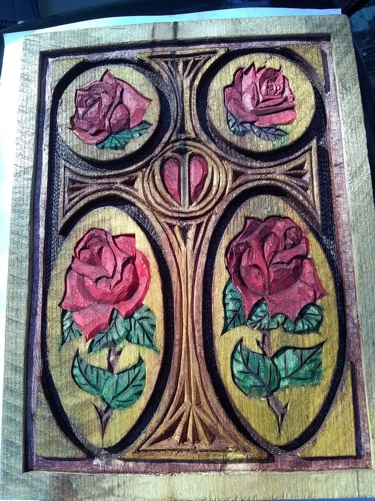 Relief carving of a cross surrounded by four red roses with green leaves and stems. A red heart carved into the center of the cross.