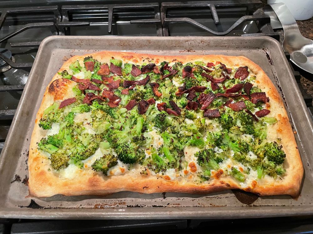 Rectangular pizza on a baking sheet. The top half of the pizza has bacon and broccoli and the bottom has just broccoli. There’s no tomato sauce. 