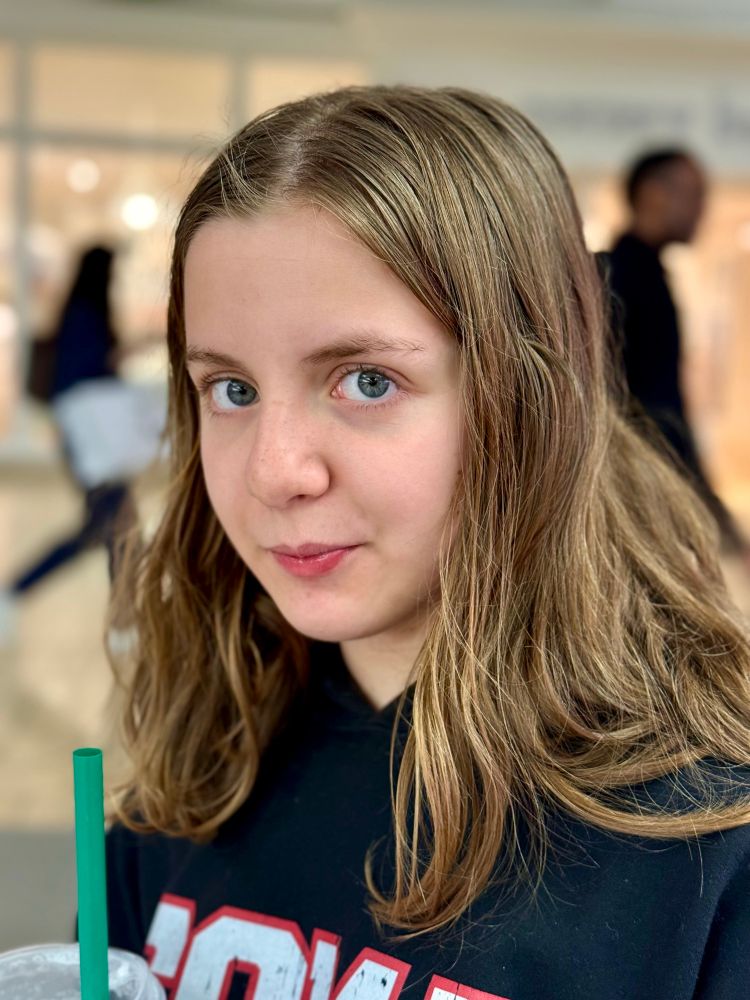 White tween girl with past shoulder length dirt blonde hair and a black hoodie with white lettering looking at the camera while holding a drink with a green straw 