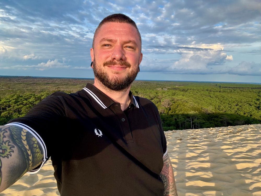 Josh standing at the top of the Grande Dune du Pilat, the tallest sand dune in Europe. It is located in La Teste-de-Buch in the Arcachon Bay area, France, 60 km southwest of Bordeaux. The sun is in his eyes, and he's possibly recovering from a very intense uphill climb to get there. He's wearing a short-sleeved black polo shirt with white accents. Has two full tattoo sleeves. The sky is intensely cloudy and sunny, and the time is near sunset, with shadows stretching far behind him. The forest canopy is visible below. 