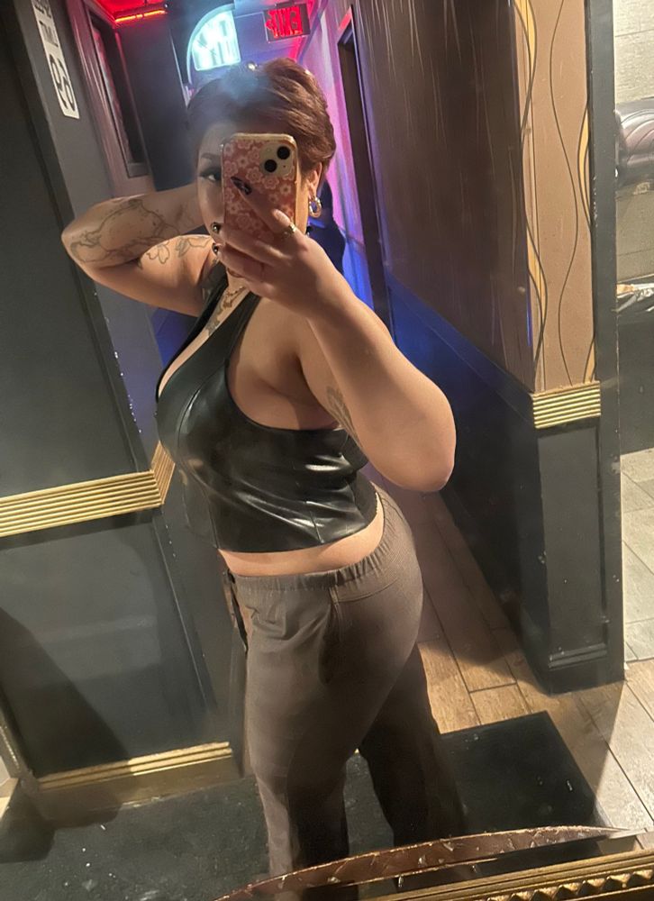 Selfie of Lena, a light skin Black person, in a hallway mirror turned to the side wearing a black halter vest and brown pants. Their short copper hair is styled in a short pixie. 
