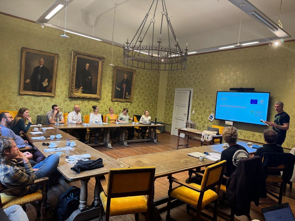 Grant Ramsey summarizes the main topics and themes of the workshop “Perspectives on Human Variation: Philosophy Meets Evolutionary Anthropology.” He stands in a historic room (Raadzaal) with ornate wallpaper, large oil portraits, and a chandelier, addressing a seated group of scientists and philosophers around a long table. 
