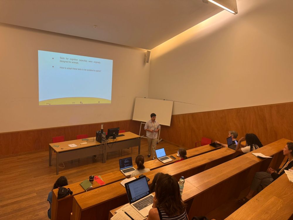 Gianmaria Dani presenting at ISHPSSB 2025. He stands in front of a projected slide in a small lecture hall with wood-paneled walls. The slide reads: 'Tests for cognitive capacities were originally designed for animals. How to adapt these tests to be suitable for plants?' Several attendees are seated in the tiered audience. 