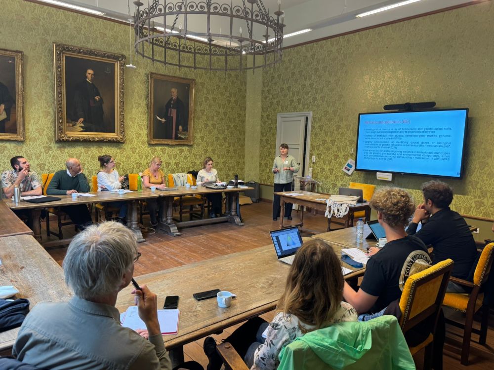 Olesya Bondarenko presents at the "Perspectives on Human Variation: Philosophy meets Evolutionary Anthropology" workshop at KU Leuven. She stands beside a screen displaying a slide titled "Behavioural genetics (BG)" while participants seated around a large wooden table listen in a historic room adorned with ornate wallpaper and large oil portraits.