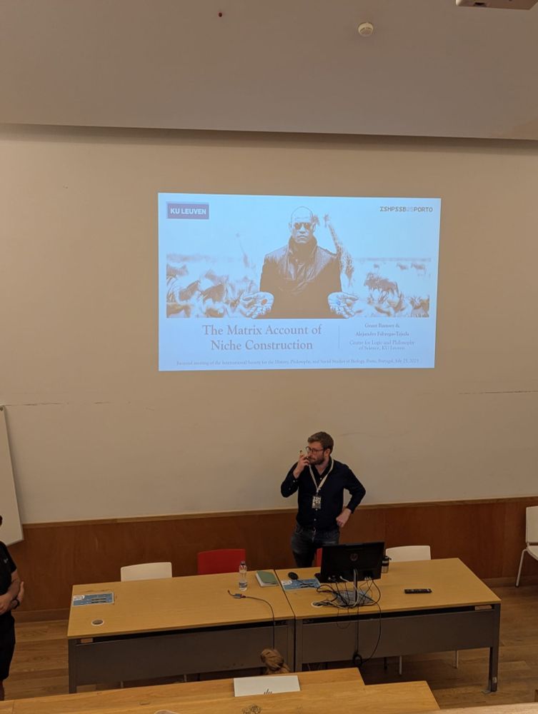 Alejandro Fábregas-Tejeda presenting at ISHPSSB 2025. He stands in front of a projected slide titled 'The Matrix Account of Niche Construction' with a stylized image of Morpheus from The Matrix.  Alejandro is in a lecture hall with wood-paneled walls and a desk with conference materials and a computer.
