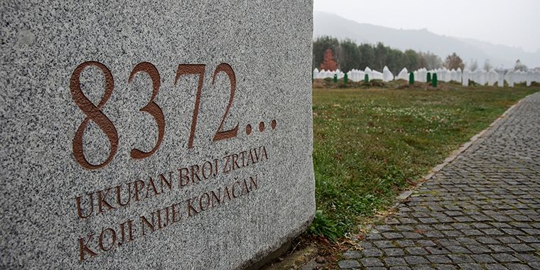 Close-up of a stone memorial at the Srebrenica genocide site in Bosnia and Herzegovina. The inscription reads "8372... Ukupan broj žrtava koji nije konačan," which translates to "8372... The total number of victims is not final." White grave markers can be seen in the background, symbolizing the lives lost.