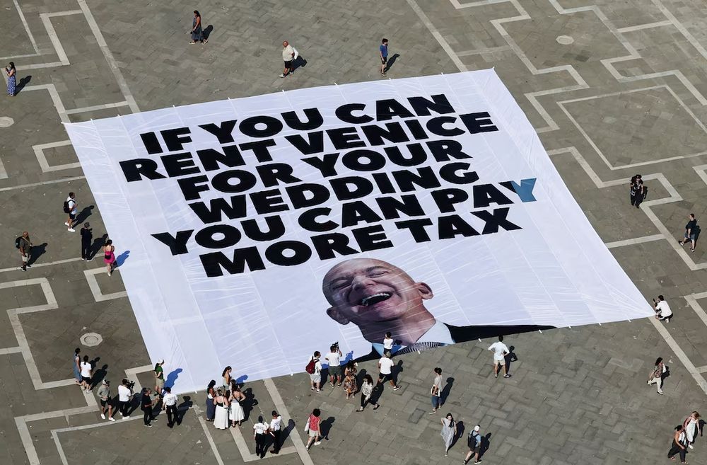 A large protest banner laid out in a public square in Venice reads: "IF YOU CAN RENT VENICE FOR YOUR WEDDING YOU CAN PAY MORE TAX" in bold black letters. The bottom of the banner features a large image of a laughing Jeff Bezos. People are gathered around and walking near the banner, some taking photos.
