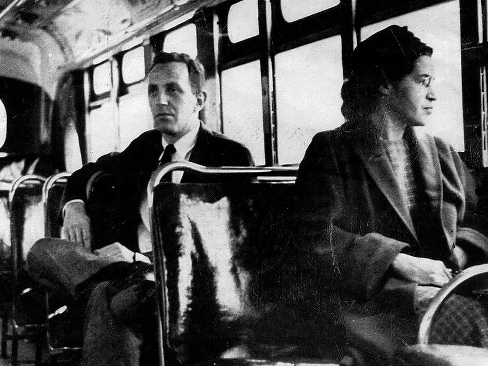 Black-and-white photo of Rosa Parks sitting on a bus in Montgomery, Alabama, in 1956. She gazes out the window with a calm yet determined expression. Behind her, a white man sits further back. This image became an enduring symbol of the U.S. civil rights movement and the fight against racial segregation.