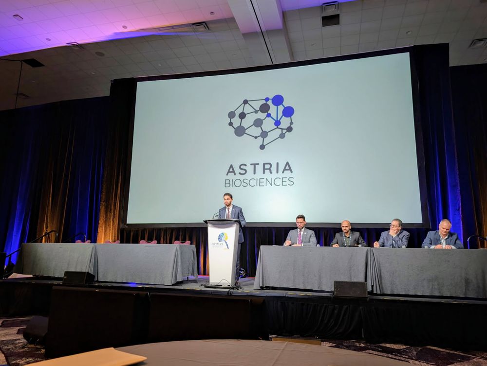 Adi Mittal, MD, presents at the SVINnovation competition. Dr. Mittal is wearing a blue suit and red tie. He is standing behind a white podium that has the SVIN 2025 logo on it. To his right are two empty tables and to his left are two tables with four judges, all of whom present as male and are wearing suits. All five individuals are in front of a projector screen that reads "Astria Biosciences" with an abstract brain logo.