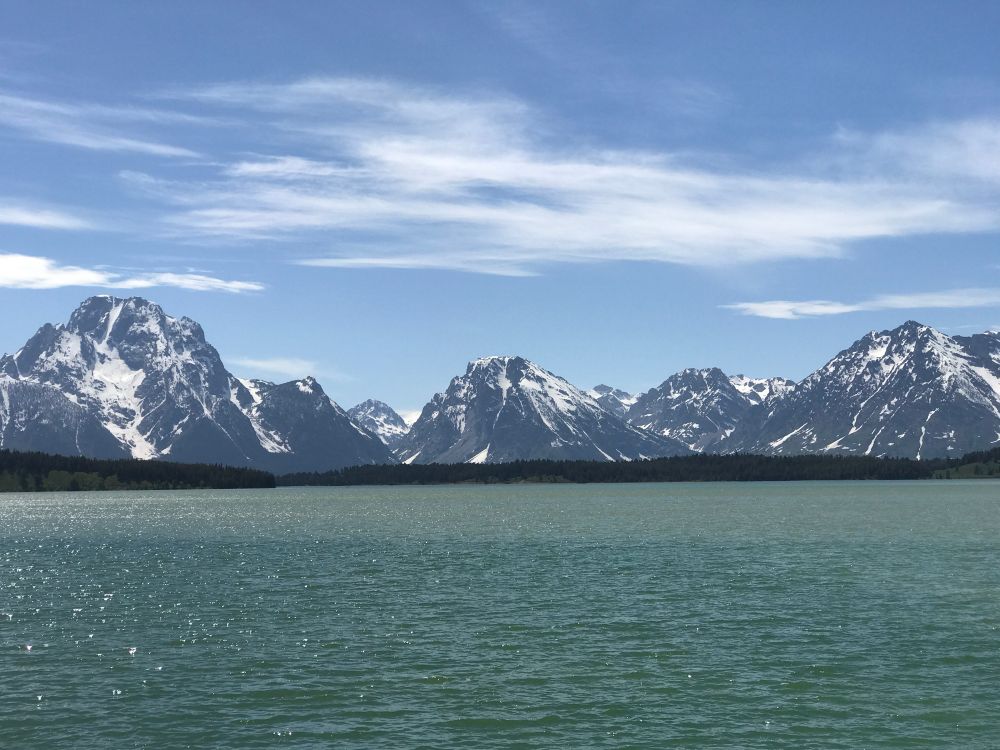 A placid, deep green lake with jagged, snow covered mountains in the distance. A blue sky with some wispy white clouds. 
