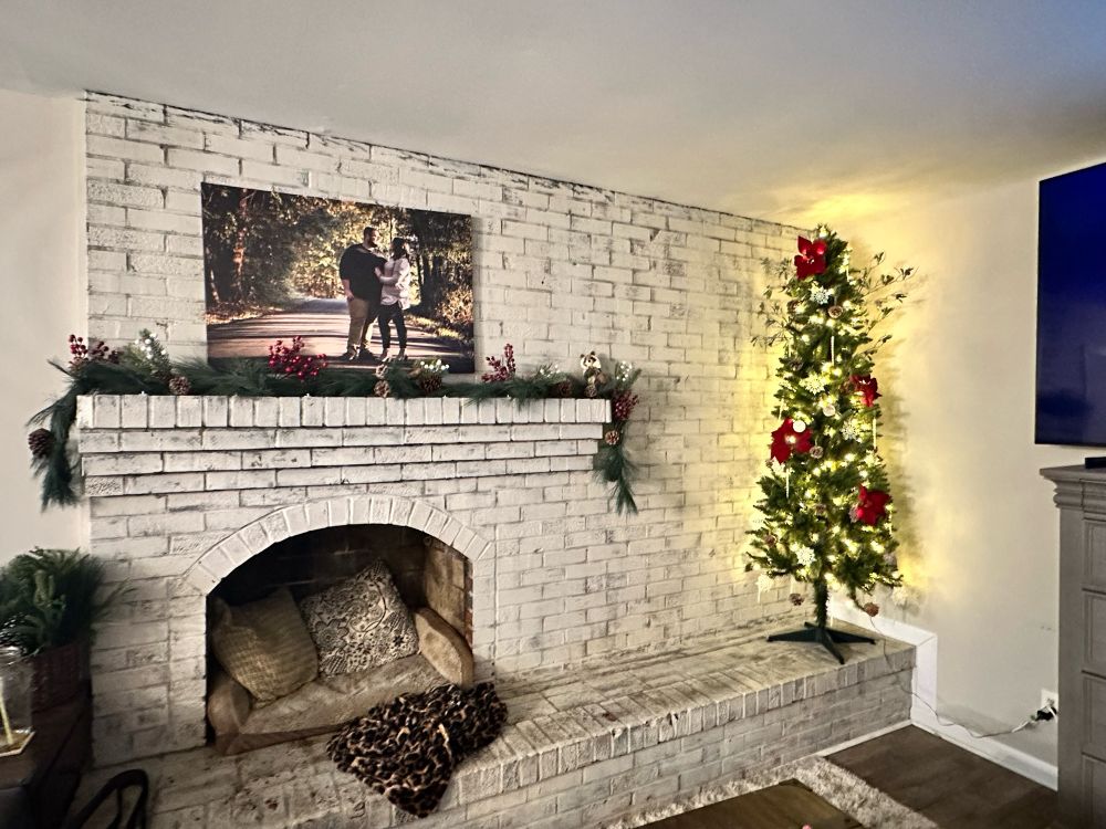 Picture of a white brick wall background with inlay fireplace, pillows inside the fireplace, a greenery garland with holly and pine cones on the mantel, below a large canvas picture of a couple standing in the middle of a road surrounded by trees. Then to the right of the fireplace is a tall Christmas tree in the corner decorated with poinsettias, pine cones and snowflakes. 