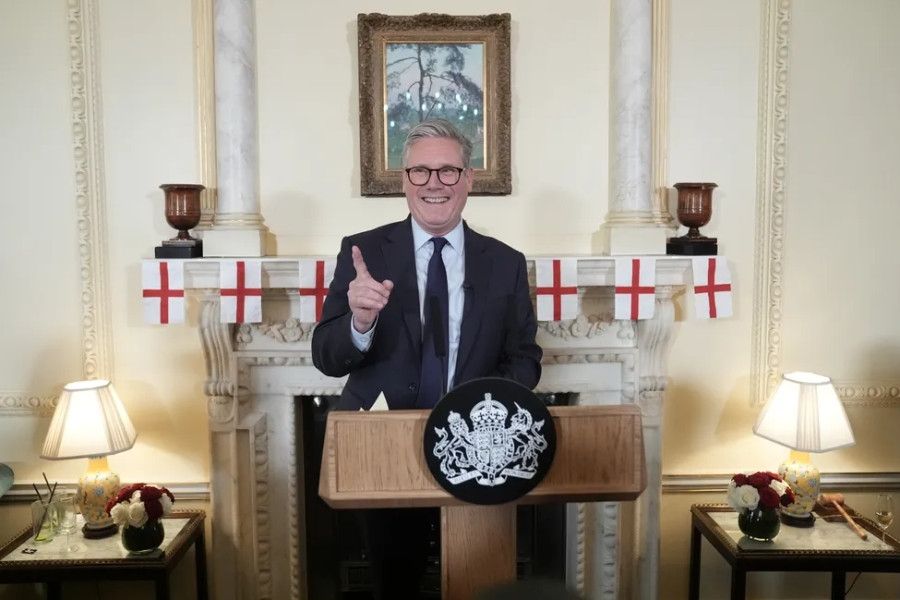 Sir Keir Starmer smiling in front of a mantle piece decked with England flags. He stands behind the lectern of PM of the UK. 