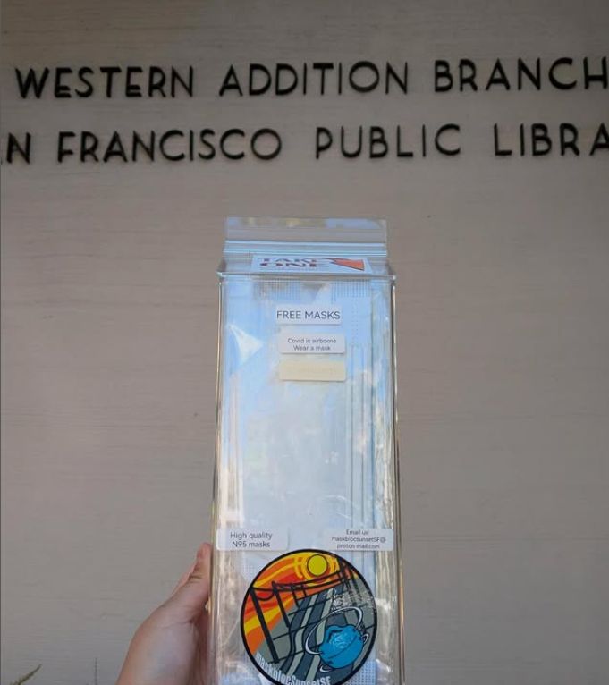 A photo of a hand by a clear plastic mask dispenser in front of a concrete wall with partially-obscured letters reading: “Western Addition Branch, San Francisco Public Library.” The mask dispenser has a sticker on top saying, “Take one” with an arrow next to it. Other stickers on the dispenser read: “Free masks.” “Covid is airborne. Wear a mask.” “High quality N95 masks.” “E-mail us at: maskblocsunsetsf @ protonmail.com”