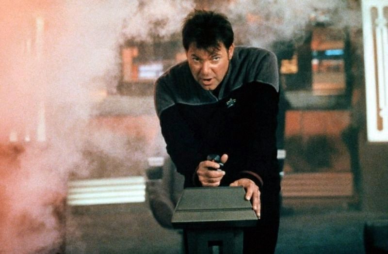 Jonathan Frakes as Will Riker in Star Trek: Insurrection. He is standing in front of standing column and firmly gripping the joystick sat atop it. Smoke surrounds him.