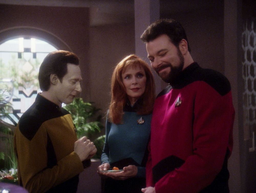 A still from the Star Trek The Next Generation episode "Starship Mine"; in which Commander Data (Brent Spiner) is looking down slightly while delivering some "small talk" to Beverly Crusher (Gates McFadden) and William Riker (Jonathan Frakes). Beverly is looking at William with a bemused expression, while William is in the midst of trying to hold in laughter. The three are standing in a beige room, with a large circular window over Data's left shoulder. There are green plants under the window.