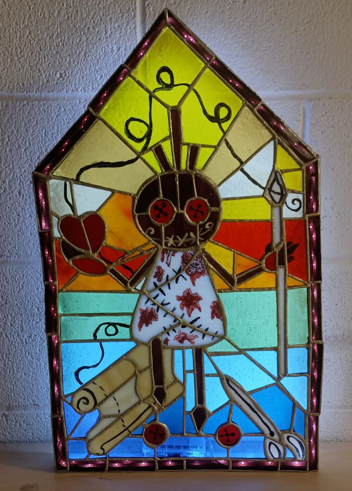 A stained glass panel of a small doll-like figure with button eyes holding a needle like a staff one one hand and a red heart in the other. By his legs is a bolt of cloth, a pair of scissors and two more loose buttons. The background is done in rainbow colors.