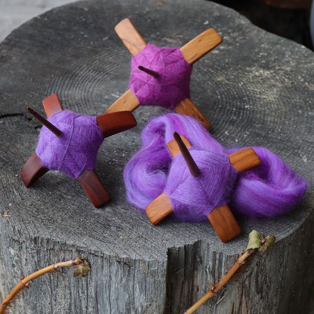 Three Turkish spindles on a grey wood stump. Pink and purple singles are spun and neatly wound and the second purple single is in progress. 