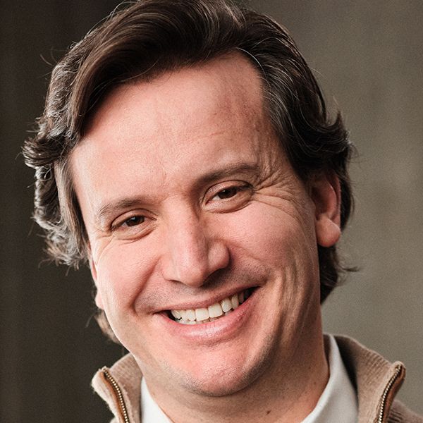 A man with short, wavy dark hair and a brown sweater smiles
