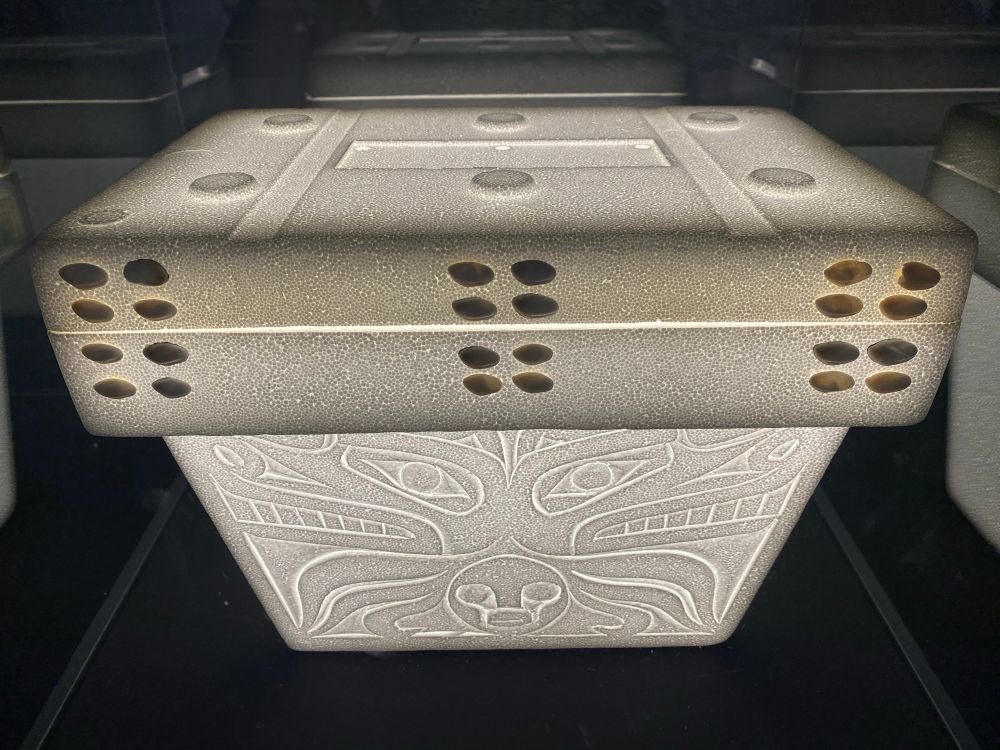 An old school styrofoam cooler with the sides carved in traditional Nuu-chah-nulth (northwest coast Indigenous) style, with operculum shells embedded in the lid. Lit from the inside, in a glass case at a gallery.