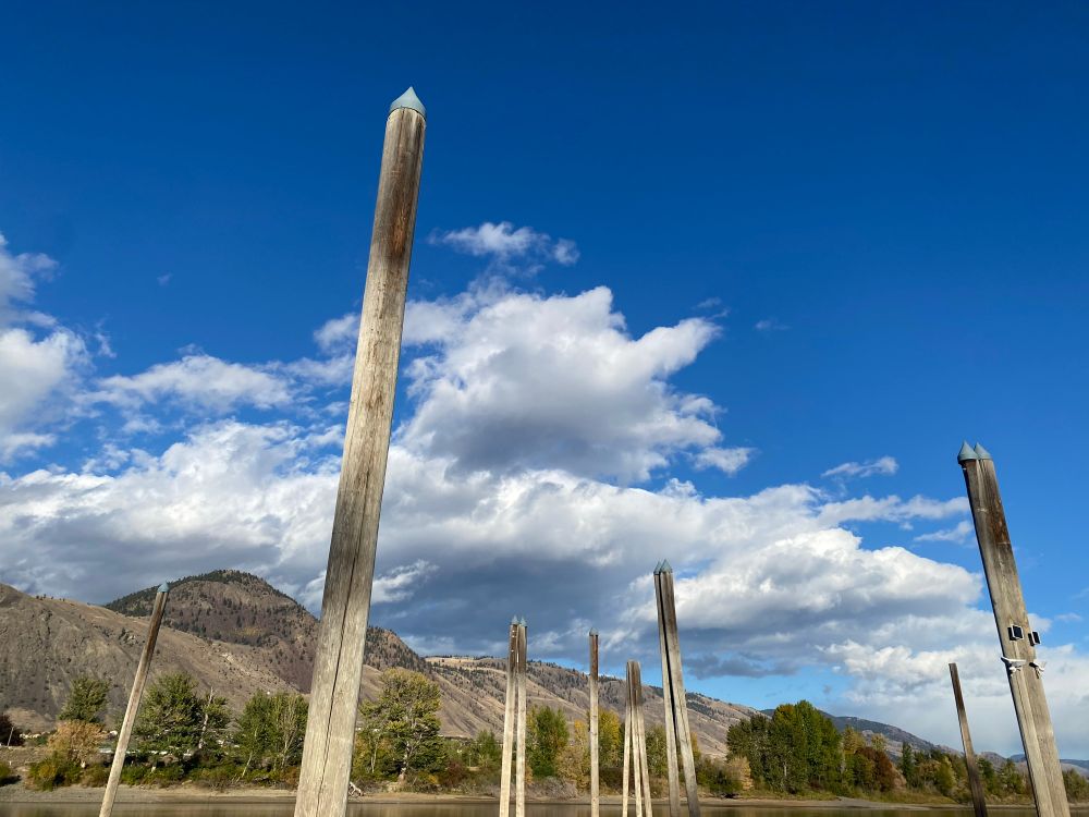 A bunch of tall poles with dull metal cones on top jutting out of the river against a background of dry grass mountains, blue sky, low white flighty clouds