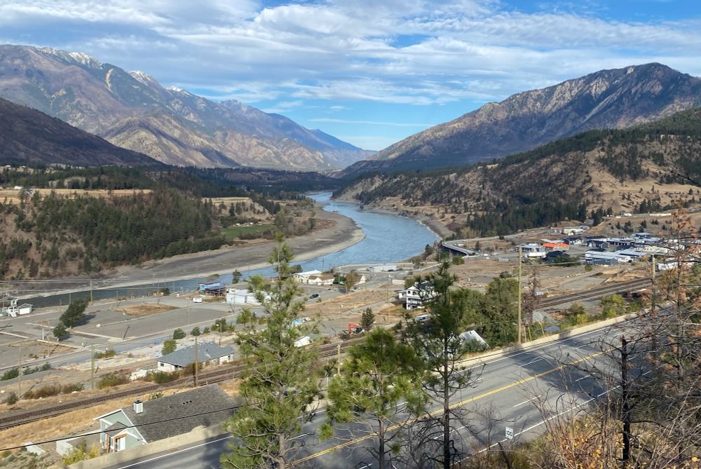 View up a large arid mountain valley with a big river chalky blue in the distance, a four lane highway at the bottom, and the village of Lytton townsite in between. A few ponderosa pines are in front of the frame.