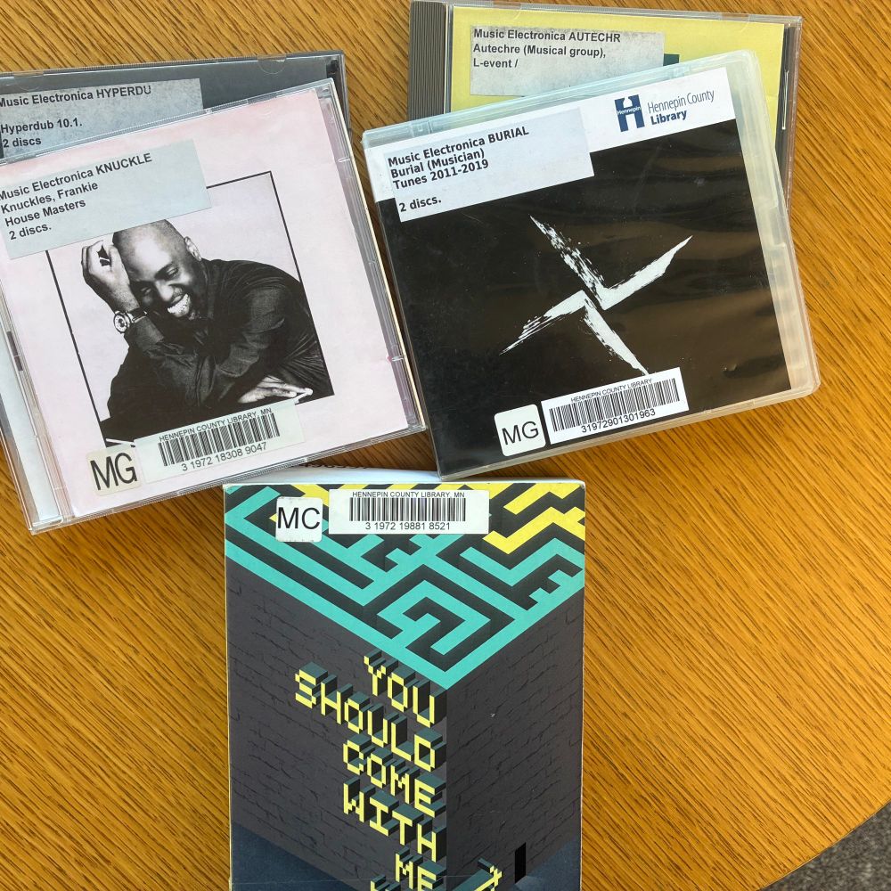 A photo of a stack of CDs in jewel cases and a book on a wood table all with Hennepin County Library barcodes. The book is You Should Come With Me Now by M. John Harrison. The CDs feature work by Burial, Frankie Knuckles, Autechre, and a collection titled Hyperdub. 