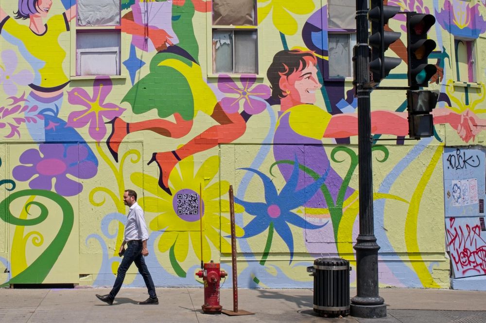 A business man in a white shirt and black paints is walking in front of a colourful mural.