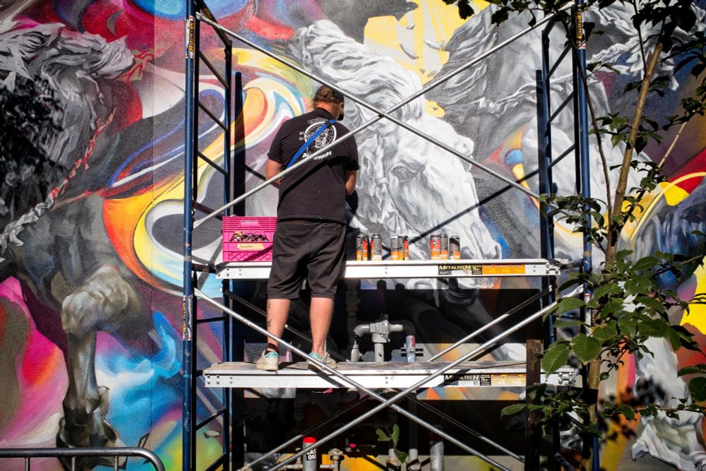 An artist is standing on scaffolding, painting a mural of horses on a wall.