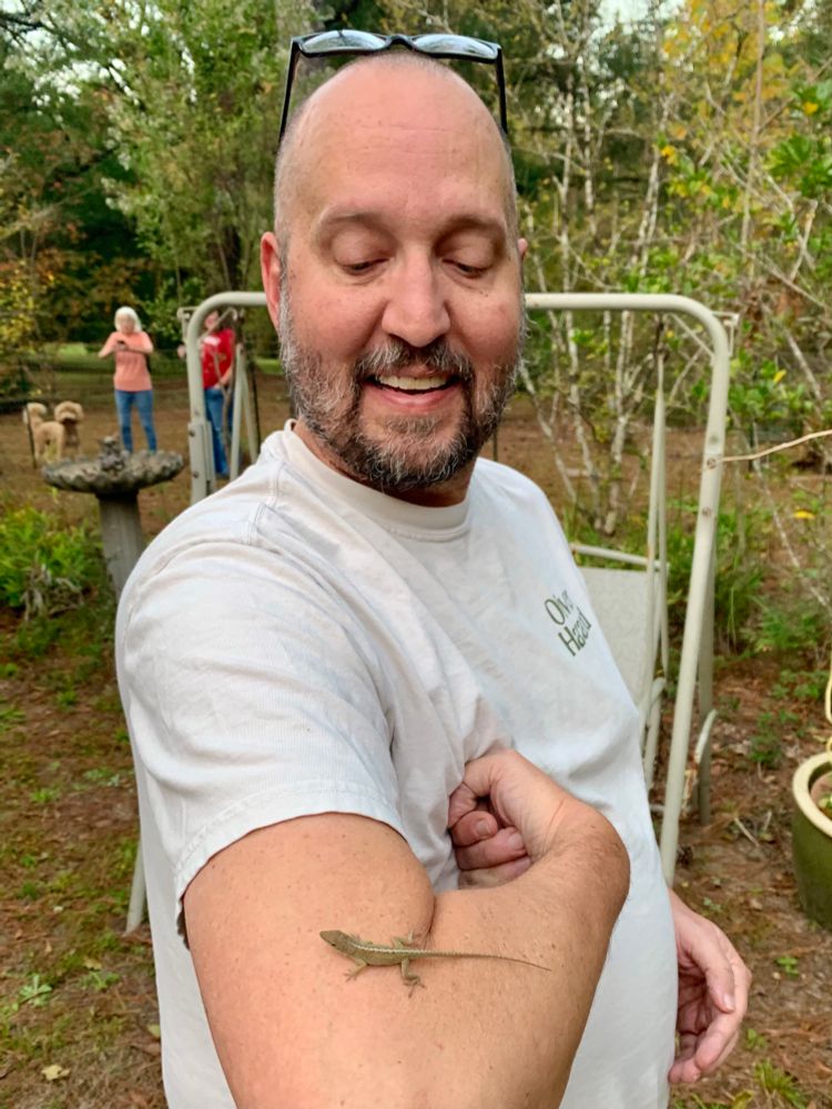 A handsome bald man with a lizard on his very nice bicep with two gorgeous ladies and a dog in the background 