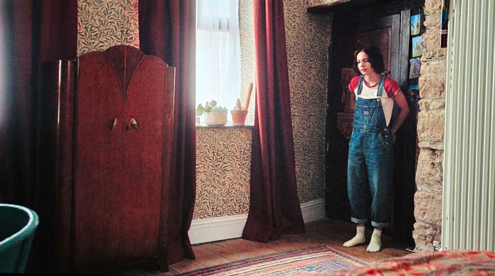 A teen girl leans against a dark wood door set in a stone wall. The adjacent wall is decorated with a swirling green and cream leaf-patterned wallpaper, long red curtains, and an Art Deco wardrobe.