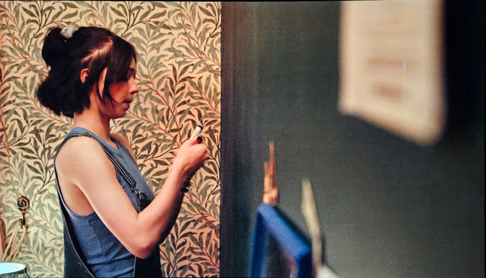 A teen girl with short dark hair stands in front of a wall decorated with green and cream leaf-patterned wallpaper.