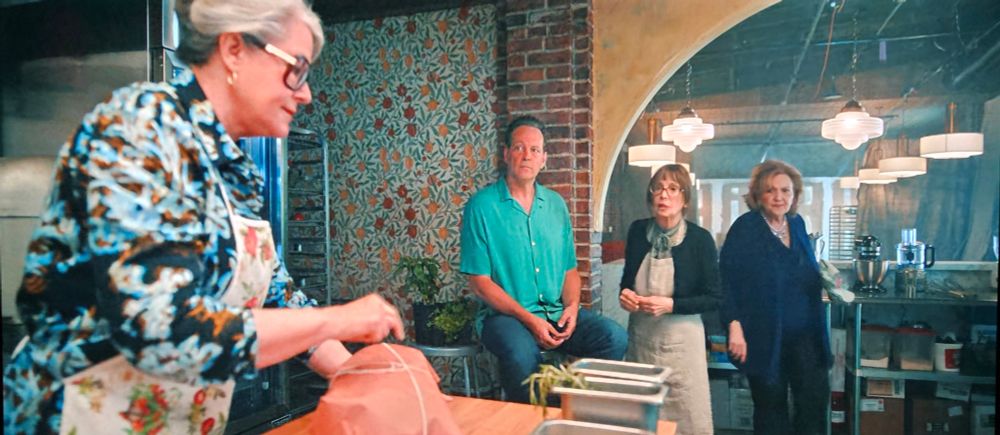 Two women and a man observe a third woman preparing a dish in a restaurant kitchen. The wall behind the man is decorated with a fruit and floral wallpaper in shades of red, yellow, green, and pale blue.