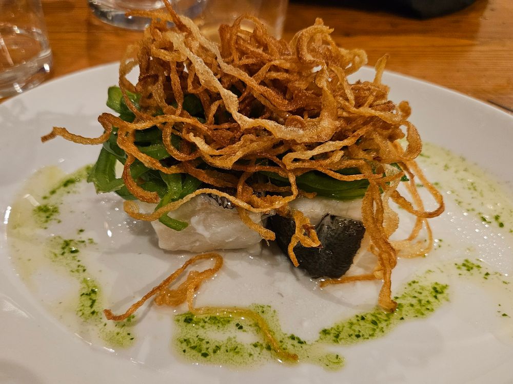A piece of white fish sits in the middle of a white plate surrounded by a herbed oil drizzle and topped with thinly sliced green peppers and crispy fried onions.