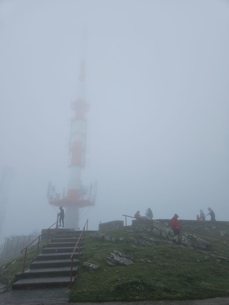 A large red and white telecommunications antenna is just barely visible through the fog at the top of a rocky summit. A few people walk around the surrounding pathways and staircases.