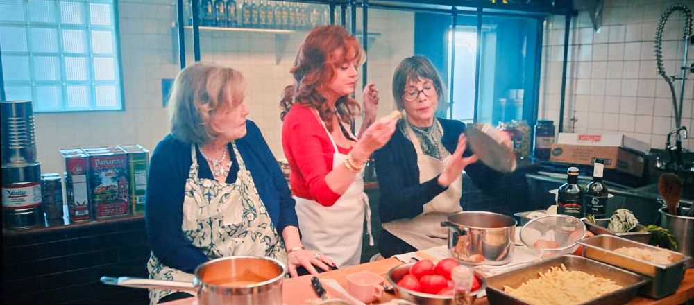 Two women sit at a counter in a restaurant kitchen while a thirs stands between them tasting some sauce. The woman on the left wears an apron decorated in a large print floral pattern in muted green shades.