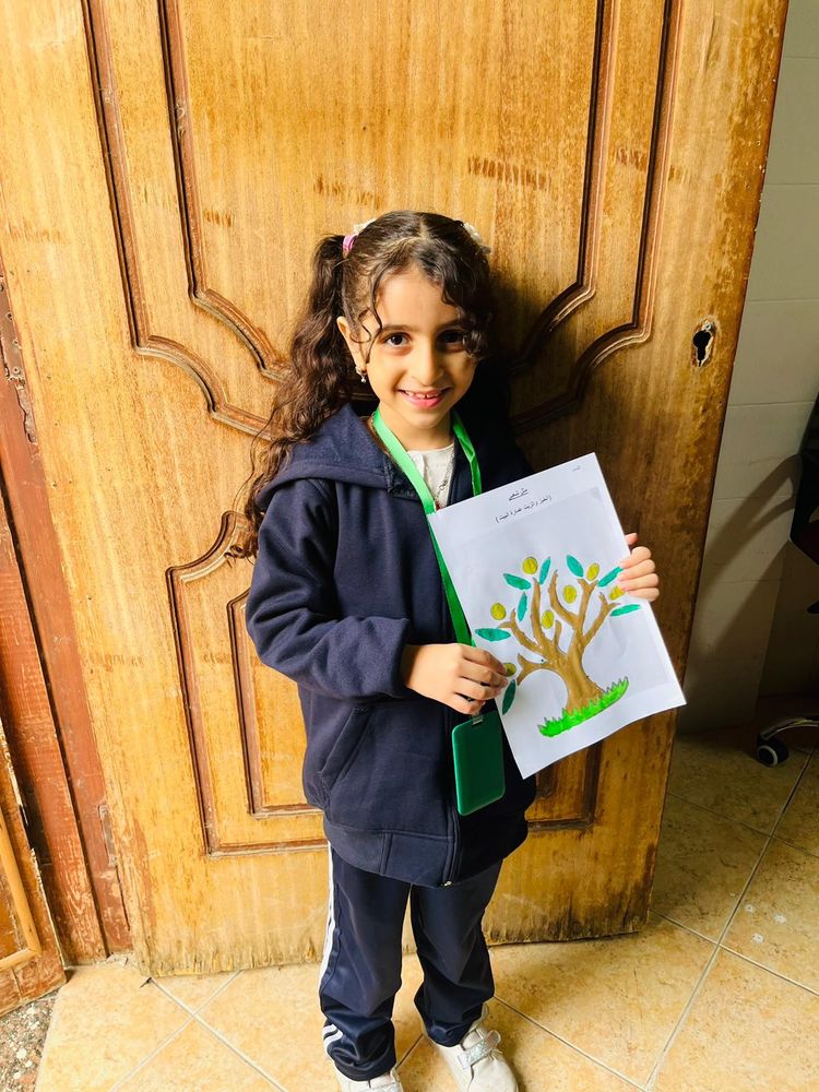 A girl in blue smiles and holds up a coloring page of an olive tree. 