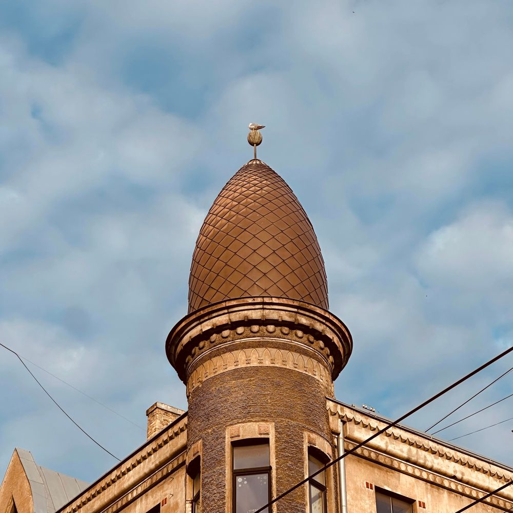A tall, rounded turret with a patterned brown roof rises from the corner of an old brick building at the intersection of Čaka and Ģertrūdes streets in Rīga, Latvia. At the very top of the turret, a seagull is perched on a golden finial. Overhead, the sky is cloudy, and some electrical wires cross in front of the building.