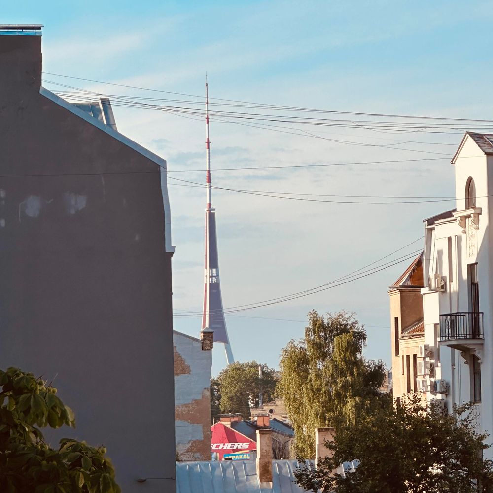 The Zaķusala TV tower stands in the background, framed between two residential buildings in Rīga. Numerous internet and cable TV wires stretch across the sky above rooftops, trees, and a few colorful building facades in the foreground, with the tall, red-and-white spire of the tower clearly visible against a bright, lightly clouded sky.