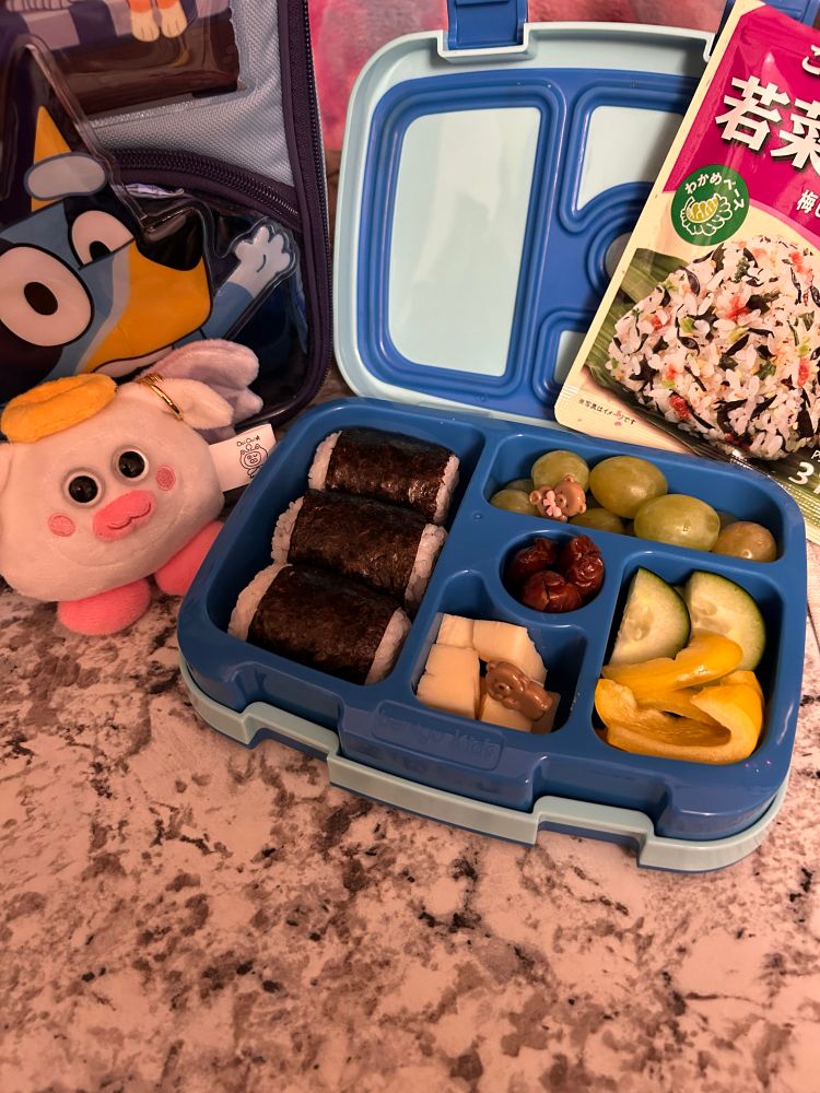 A blue bento lunch box filled with fruits and vegetables, and an angel ouioui plush, by a Bluey lunchbox. 