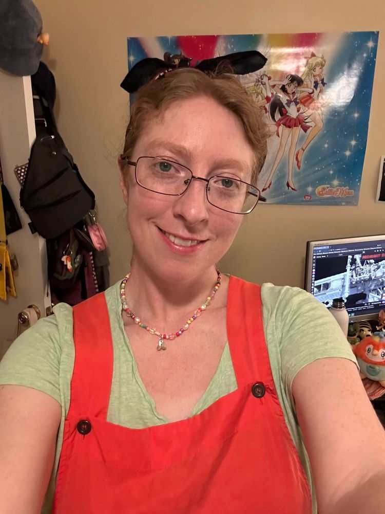 A selfie of a femme looking person, wearing a green shirt and red cotton shortalls.