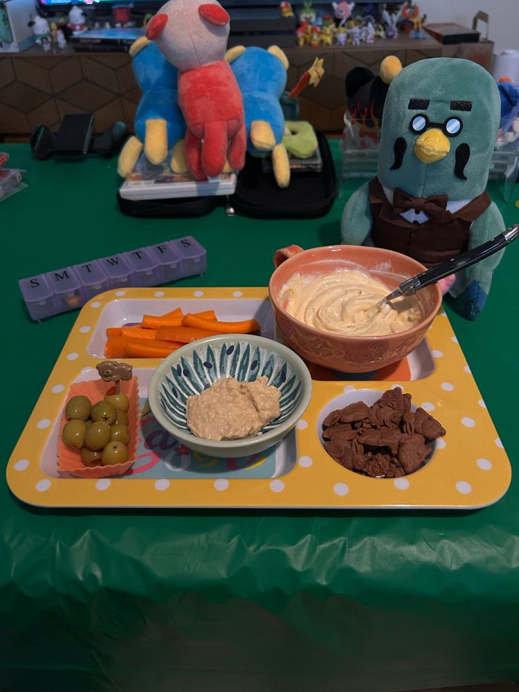 Photo of a meal on a divided plate, showing carrot sticks, chocolate bunny crackers, olives, hummus, and yogurt. 