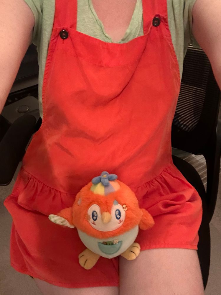 A selfie of a femme person wearing red cotton shortalls, with a cute bird plushie in their lap.  