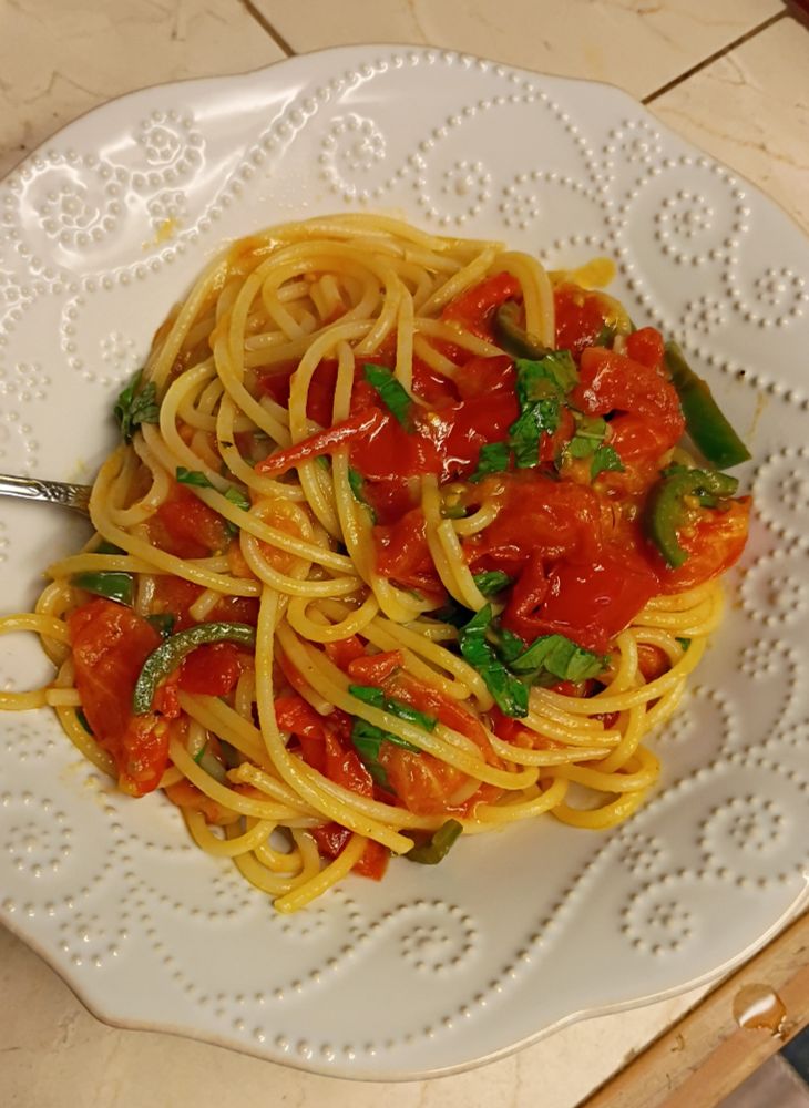 A white bowl with spaghetti noodles tossed with sauteed tomatoes, a pepper, and sliced fresh basil.