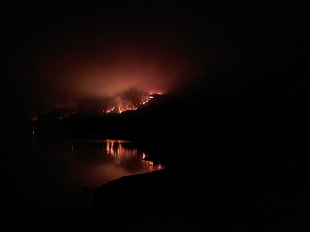 A nighttime photo of a fiery hillside reflected in water. It's extra stunning because it's a mostly black photo with a line of fire and glow all that's visible on the hill and a mostly clear reflection of that in the water.