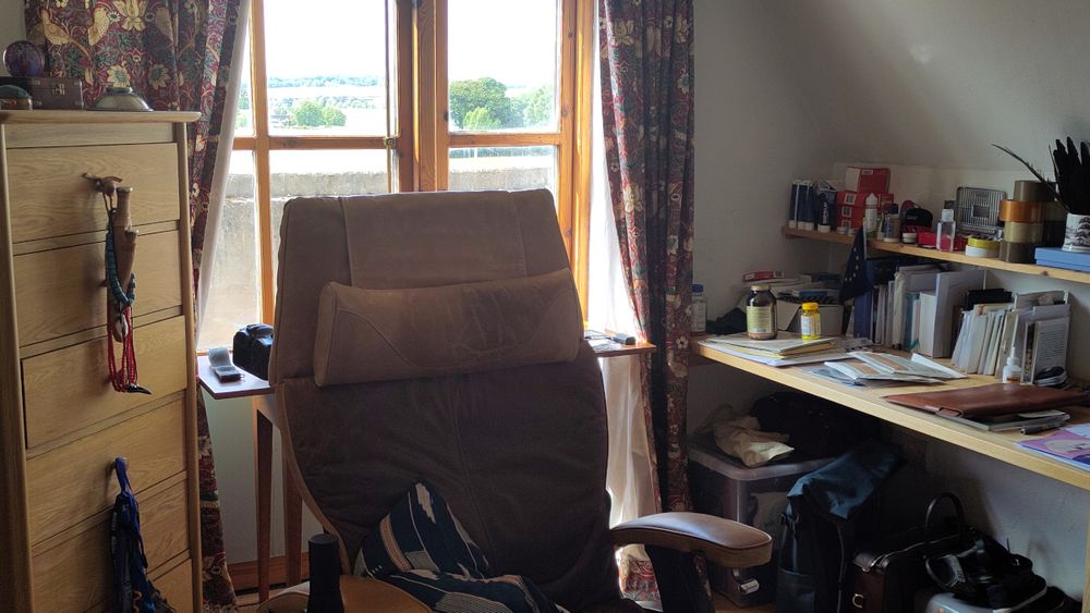 An ergonomic chair sits welcoming in front of two desks and a window with a view of trees receding into the distance