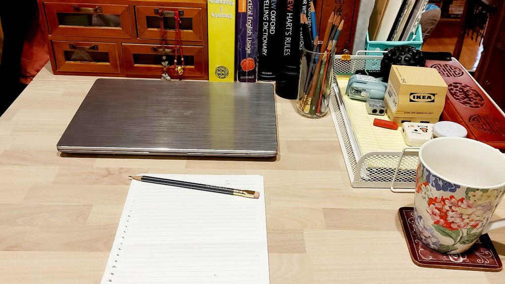 A close-up of a writer's desk, with laptop, mug and a few sheets of blank paper. But who could it be?