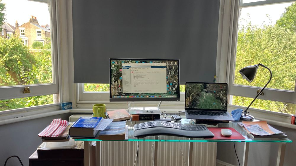 A blind is drawn in the window behind a glass desk with a large screen perched on a thesaurus and a laptop perched on a dictionary