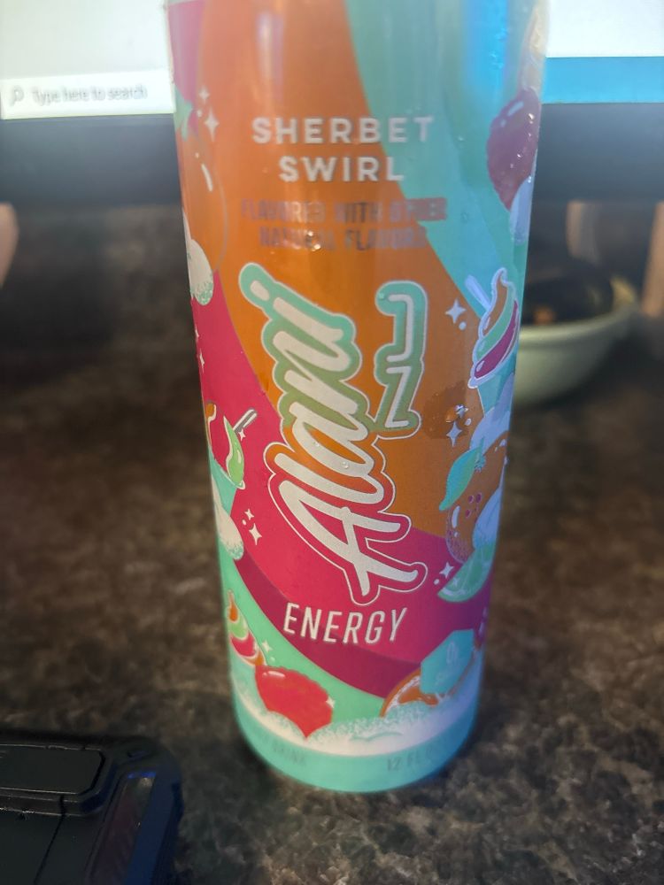 A can of Alani Nu brand energy drink in the flavor Sherbet Swirl sits on a desk. The can is brightly colored with art of fruits and swirls of sherbet.